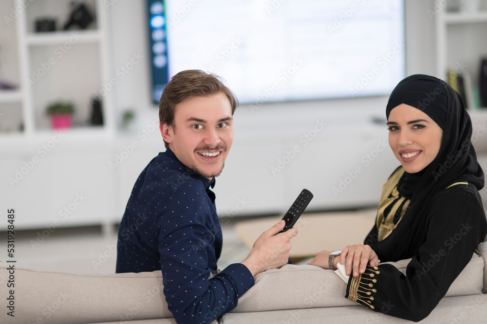 Fototapeta premium Young muslim couple woman wearing islamic hijab clothes sitting on sofa watching TV together during the month of Ramadan at modern home