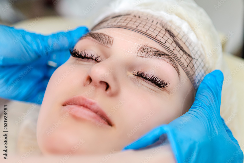 Professional Tattoo Markup with Transparent Facial Ruler On Eyebrows of Young Caucasian Woman During a Process of Permanent make up in Salon. Professional Tattoo Markup with Transparent Facial Ruler On Eyebrows of Young Caucasian Woman During a Process of Permanent make up in Salon.