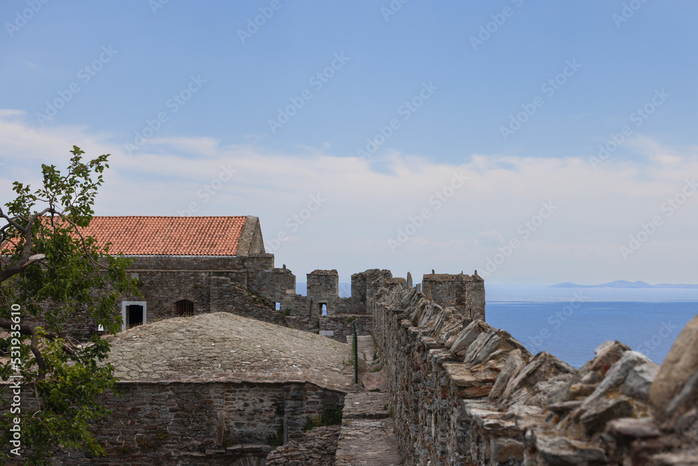 Battlements of castle walls, horizon consisting of sea and mountain range silhouette, tiled roof, and green tree. Kavala, Greece.