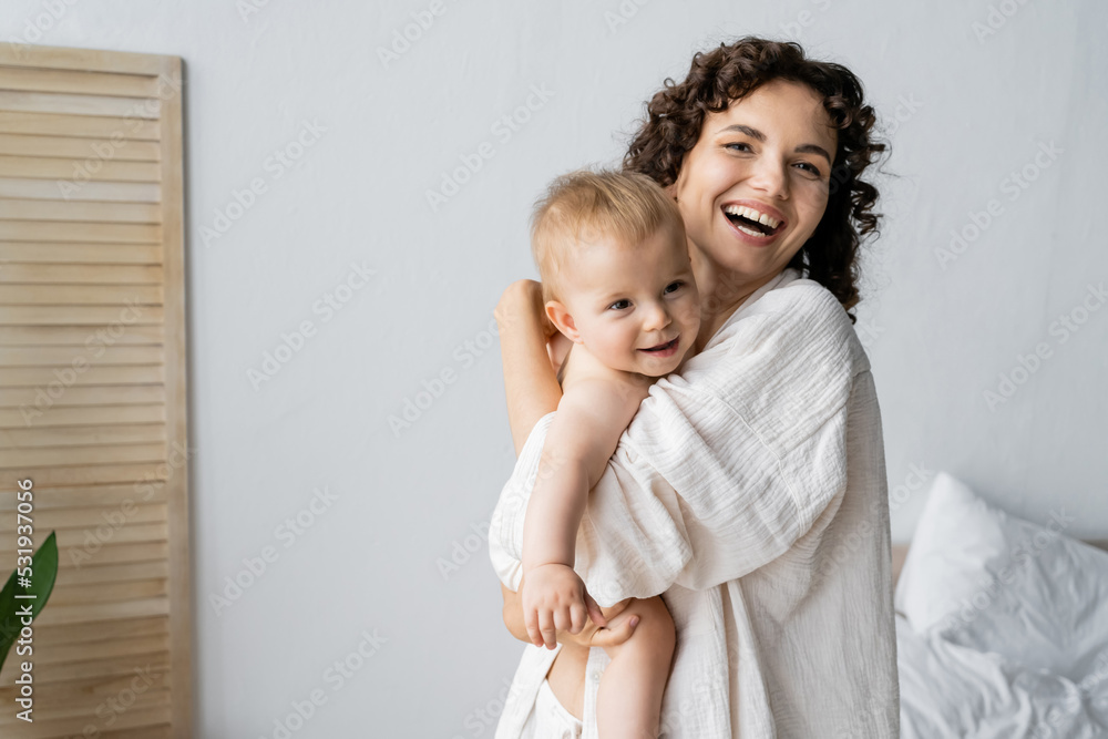 Happy woman looking at camera while hugging baby daughter in bedroom. Happy woman looking at camera while hugging baby daughter in bedroom.