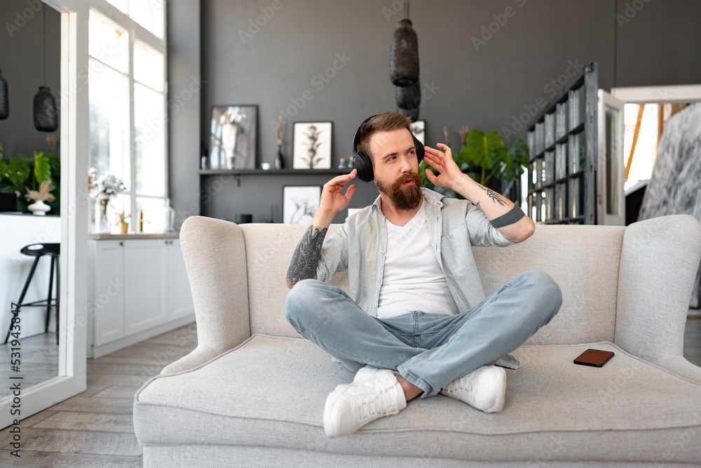 Fototapeta premium Positive bearded man listening music with headphones while sitting on couch at home