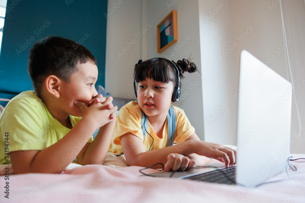 Asian kid sister using headphones and laptop study online with young brother video call to teacher at bedroom. Asian kid sister using headphones and laptop study online with young brother video call to teacher at bedroom.