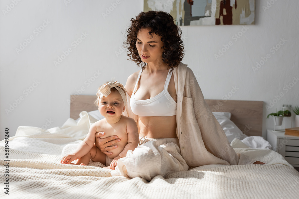 curly mother in loungewear with crop top sitting on bed with infant daughter in headband. curly mother in loungewear with crop top sitting on bed with infant daughter in headband.