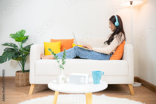 A teenage girl is happily watching something on a laptop computer in the living room of her home. Listening music by headphone by sitting on the white sofa.