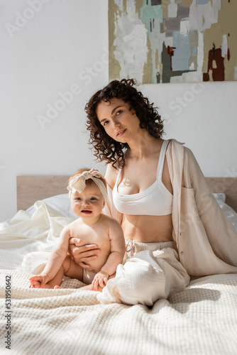 curly woman in loungewear with crop top sitting on bed with infant daughter in headband.