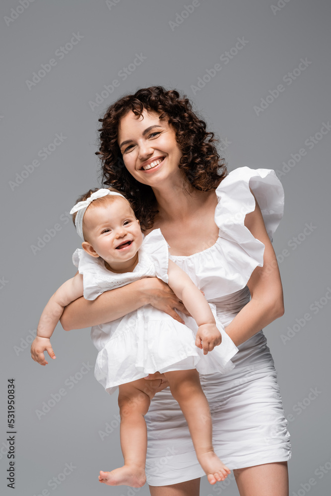 Brunette woman looking at camera while holding baby in dress isolated on grey. Brunette woman looking at camera while holding baby in dress isolated on grey.