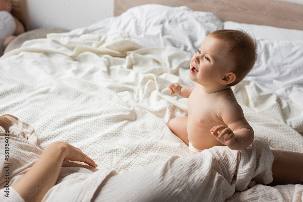 happy infant baby sitting on bed and looking at mother resting in bedroom. happy infant baby sitting on bed and looking at mother resting in bedroom.