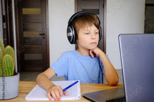 a schoolboy boy sits in front of a monitor in headphones, does his homework, listens to an online lesson, learns new school material.