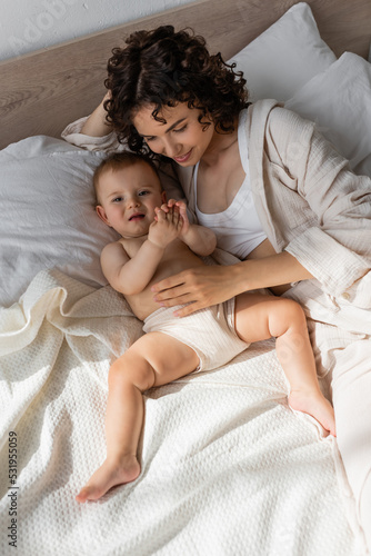 high angle view of pleased curly mother in loungewear lying with baby girl on bed.