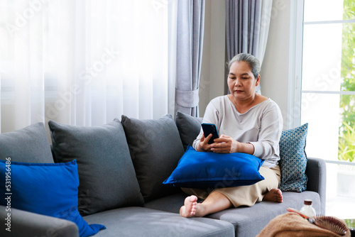 Alone retired asian older woman sitting on sofa and talk to her family by mobile phone is using internet at living room, relax time at home.