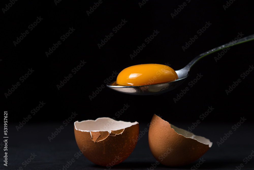 Egg yolk in a spoon next to the shell on a black background. Broken chicken egg. - obrazy, fototapety, plakaty 