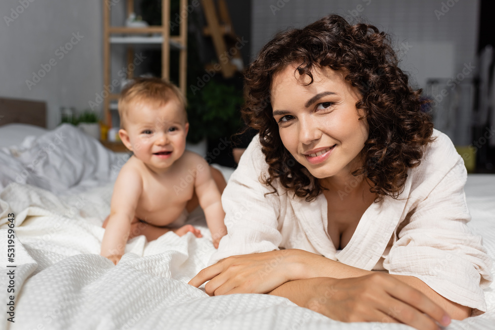 cheerful mother with curly hair looking at camera joyful baby daughter on bed. cheerful mother with curly hair looking at camera joyful baby daughter on bed.