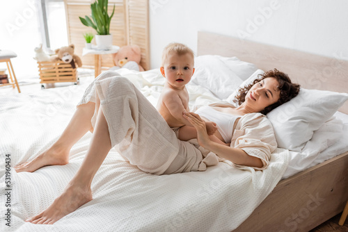 happy woman in loungewear lying on bed and looking at curious infant daughter. photo