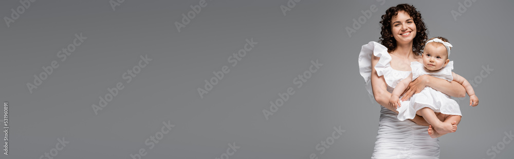 Curly mom in white dress holding baby girl isolated on grey, banner. Curly mom in white dress holding baby girl isolated on grey, banner.