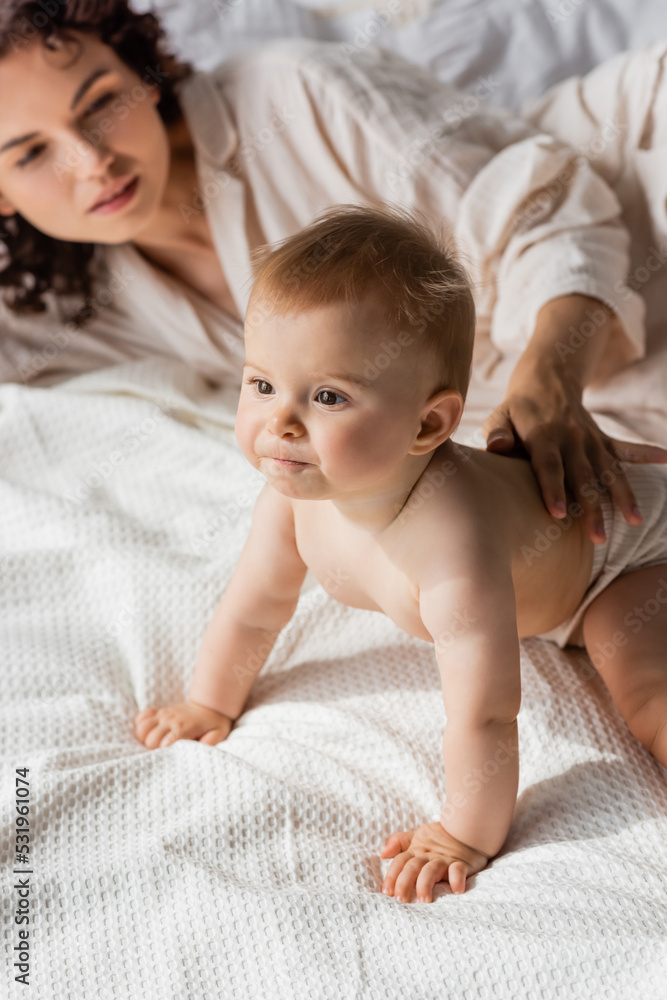 curly mother touching back of infant daughter crawling on bed. curly mother touching back of infant daughter crawling on bed.