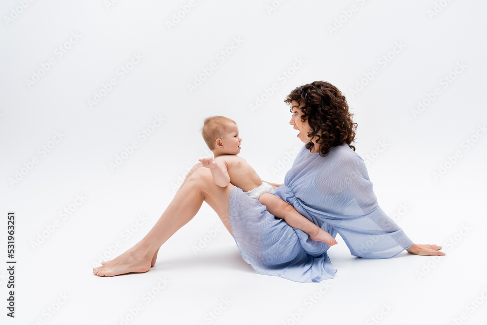 Side view of shocked mother in dress looking at baby girl in panties on white background. Side view of shocked mother in dress looking at baby girl in panties on white background.