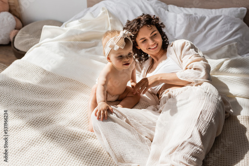happy mother with curly hair lying on bed near cute infant daughter in headband.