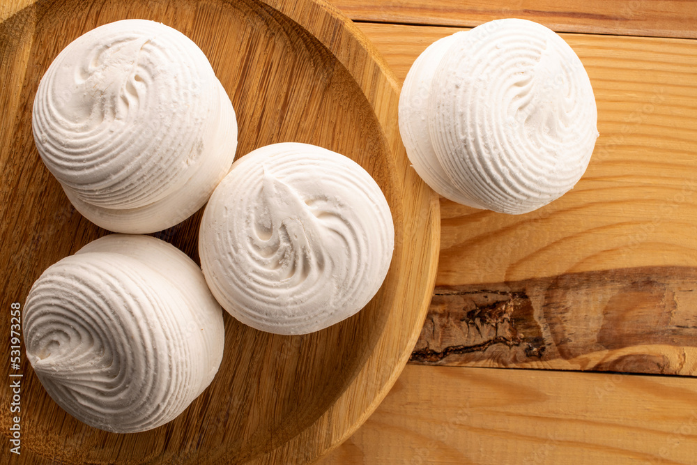 Several white sweet marshmallows on a round bamboo tray on a wooden table, close-up, top view. Several white sweet marshmallows on a round bamboo tray on a wooden table, close-up, top view.