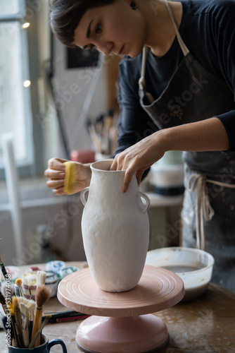 Young woman works with big vase using special tool to make pitcher smooth shape. Brown-haired craftswoman with concerned expression puts big effort into clay vase to send to exhibition in museum