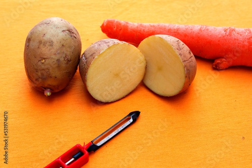 Papas y zanahoria caseras de la vida cotidiana sobre la mesa con el instrumento de cocina del pelapapas de acero y mango rojo, forman un diseño original culinario con fondo naranja