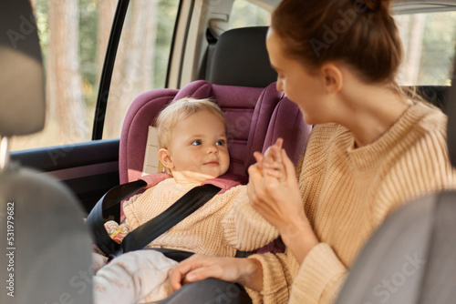 Portrait of smiling Caucasian mother in beige sweater and her little daughter in baby seat traveling by car, playing together during their journey, expressing happiness.