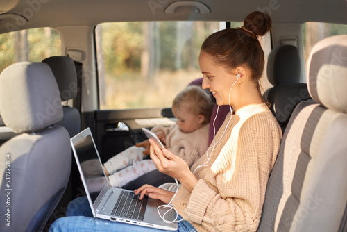 Profile portrait of dark haired smiling satisfied woman working on laptop while sitting with her baby daughter in safety chair on backseat of car, young adult girl using cell phone and listening music © sementsova321
