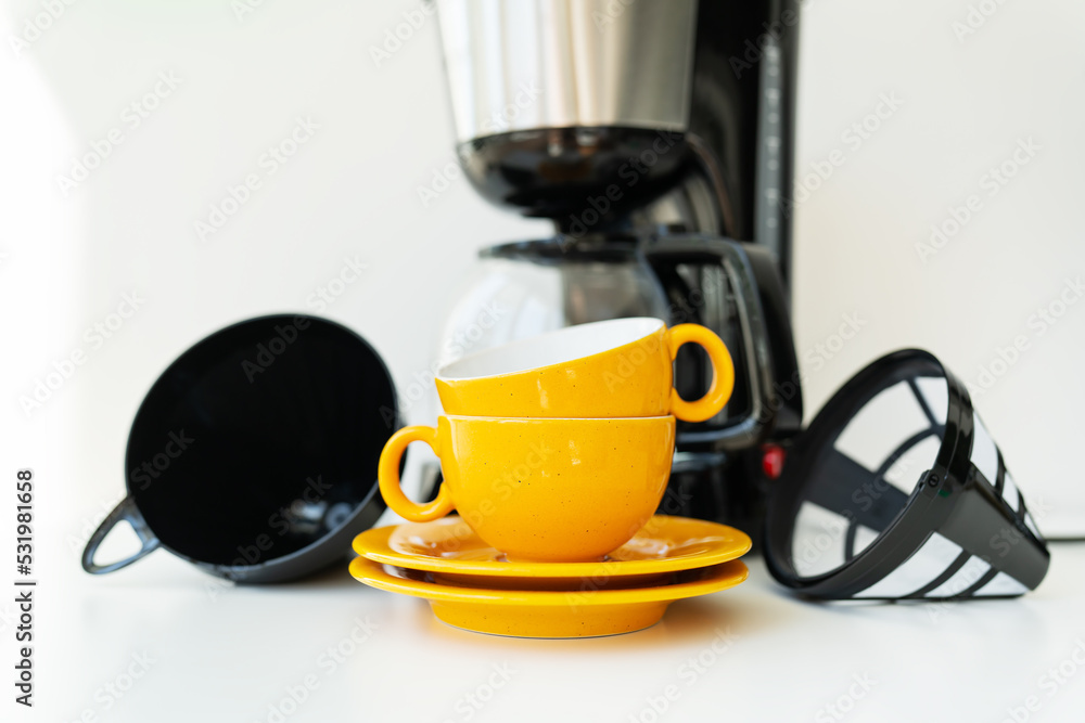 An automatic drip coffee maker stands with a yellow cup on a white table. Black modern drip coffee pot. Electric kitchen small household appliances. Appliances. An automatic drip coffee maker stands with a yellow cup on a white table. Black modern drip coffee pot. Electric kitchen small household appliances. Appliances.