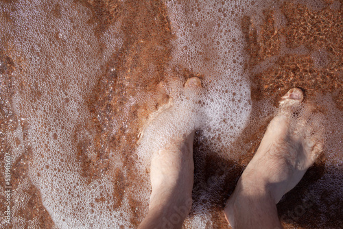 Top view of men s legs in the sea. A man is standing on the sand. An ocean wave washes the shore. Summer holidays.