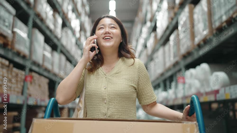 Happy Asian woman pushing shopping cart while talking on phone at large warehouse retail store. Rack of furniture and home accessories store. Interior of cargo logistic concept. Happy Asian woman pushing shopping cart while talking on phone at large warehouse retail store. Rack of furniture and home accessories store. Interior of cargo logistic concept.