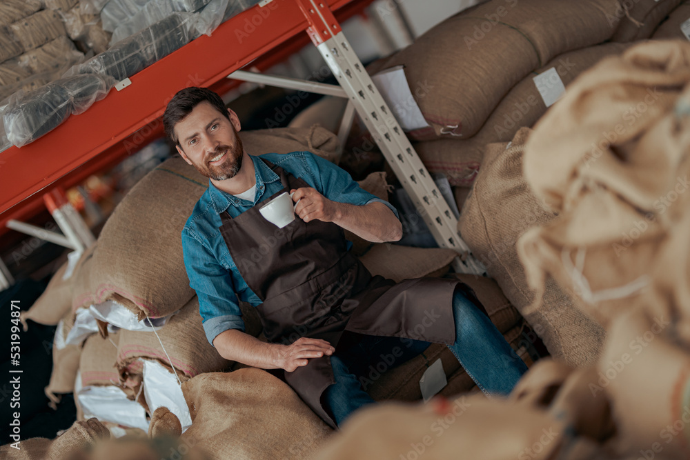 Smiling Business owner with cup of fresh coffee sitting on bags with roasted coffee beans Smiling Business owner with cup of fresh coffee sitting on bags with roasted coffee beans