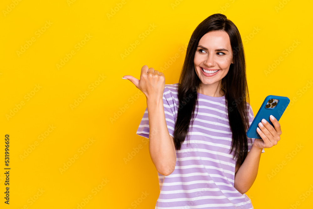 Photo of positive lovely cute lady toothy smile trendy striped clothes arm hold telephone finger direct empty space isolated on yellow color background Photo of positive lovely cute lady toothy smile trendy striped clothes arm hold telephone finger direct empty space isolated on yellow color background