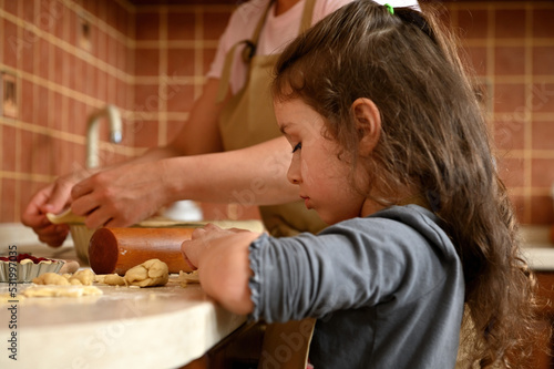 Beautiful European little girl with long hair, using rolling pin, rolling out the dough, cooking a delicious homemade cherry pie with her mother, standing by a kitchen countertop in the home interior photo