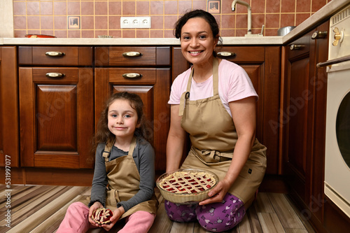 Lovely charming beautiful tender gentle mom and her adorable kid, a little daughter holding in hands pies with sweet cherry berries, smiling at camera, sitting together on the floor of home kitchen photo