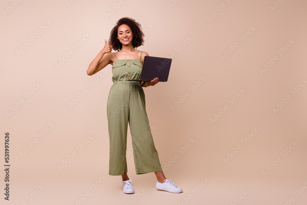 Full size photo of cool brunette woman hold laptop thumb up wear khaki overall isolated on beige color background Full size photo of cool brunette woman hold laptop thumb up wear khaki overall isolated on beige color background