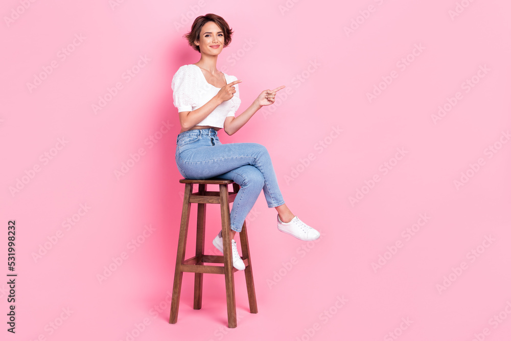 Full length photo of pretty shiny lady wear white crop top sitting wooden chair pointing empty space isolated pink color background Full length photo of pretty shiny lady wear white crop top sitting wooden chair pointing empty space isolated pink color background