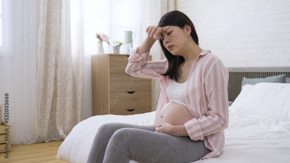 portrait asian expectant mom is feeling dizzy and rubbing her brows with discomfort on bed in the bedroom at home. portrait asian expectant mom is feeling dizzy and rubbing her brows with discomfort on bed in the bedroom at home.
