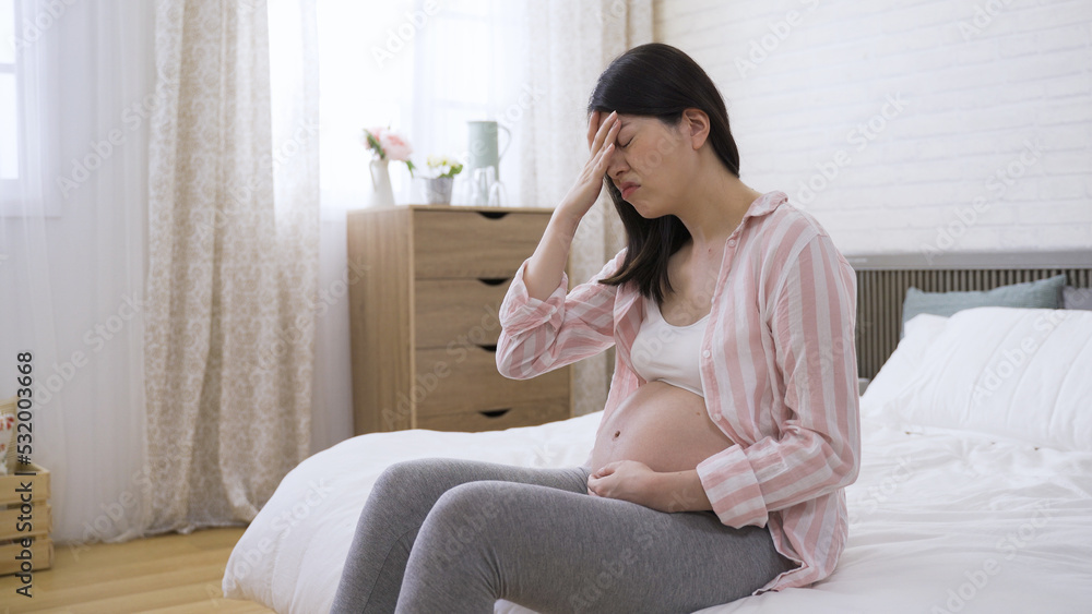 chinese pregnant lady sitting on bed and touching her forehead with painful look is suffering dizziness at bright home interior. chinese pregnant lady sitting on bed and touching her forehead with painful look is suffering dizziness at bright home interior.