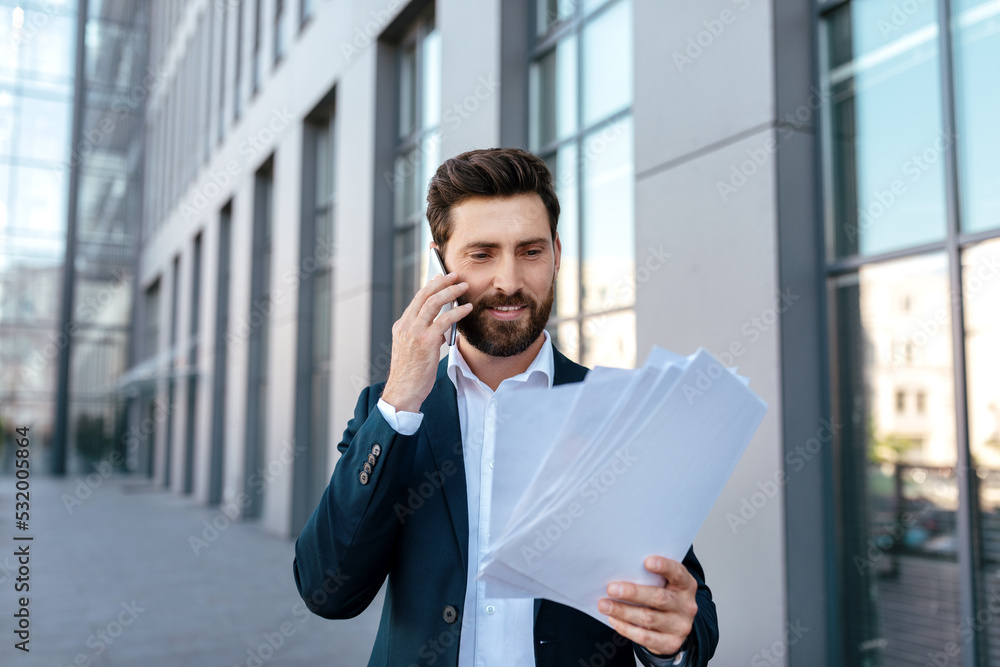 Busy confident attractive young caucasian man with beard calls by smartphone, works with documents Busy confident attractive young caucasian man with beard calls by smartphone, works with documents