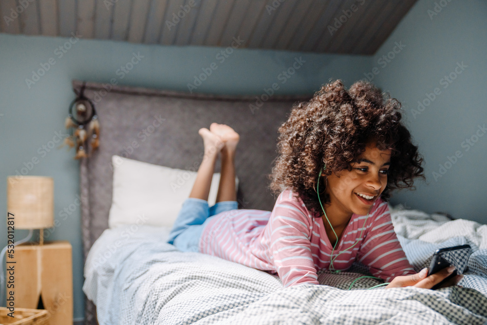 Black girl in earphones using mobile phone while resting on bed Black girl in earphones using mobile phone while resting on bed