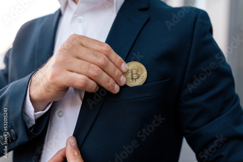 Confident young caucasian guy ceo manager in suit puts bitcoin coin in pocket near modern outdoor, close up