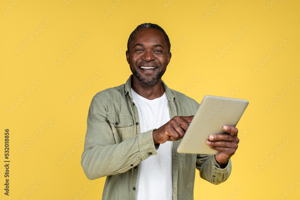 Happy surprised mature african american male in casual typing on tablet, isolated on yellow background, studio Happy surprised mature african american male in casual typing on tablet, isolated on yellow background, studio
