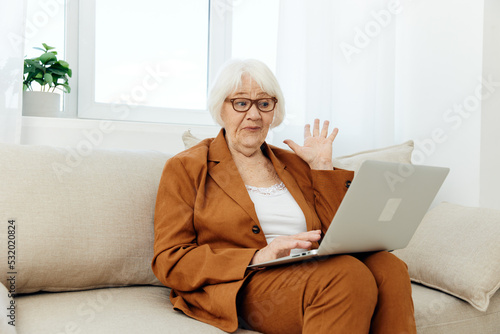 a very surprised elderly woman is sitting looking at a laptop on a sofa in a bright room near the window with glasses on her face and looks shocked with her hand raised up © Tatiana
