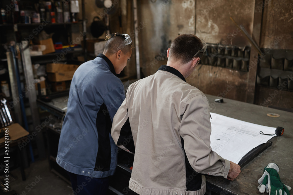Back view of two workers looking at blueprints and plans in industrial factory workshop Back view of two workers looking at blueprints and plans in industrial factory workshop