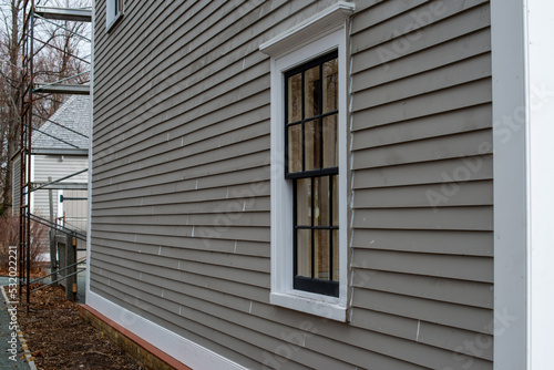 Old tan colored house with white trim and black wood window frame spacers. The glass in the window is wavy and reflects the sun. The exterior wall is made of vintage beige cape cod clapboard siding.