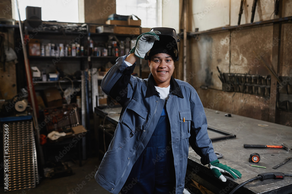 Waist up portrait of multiethnic female welder smiling at camera in industrial factory workshop, copy space
