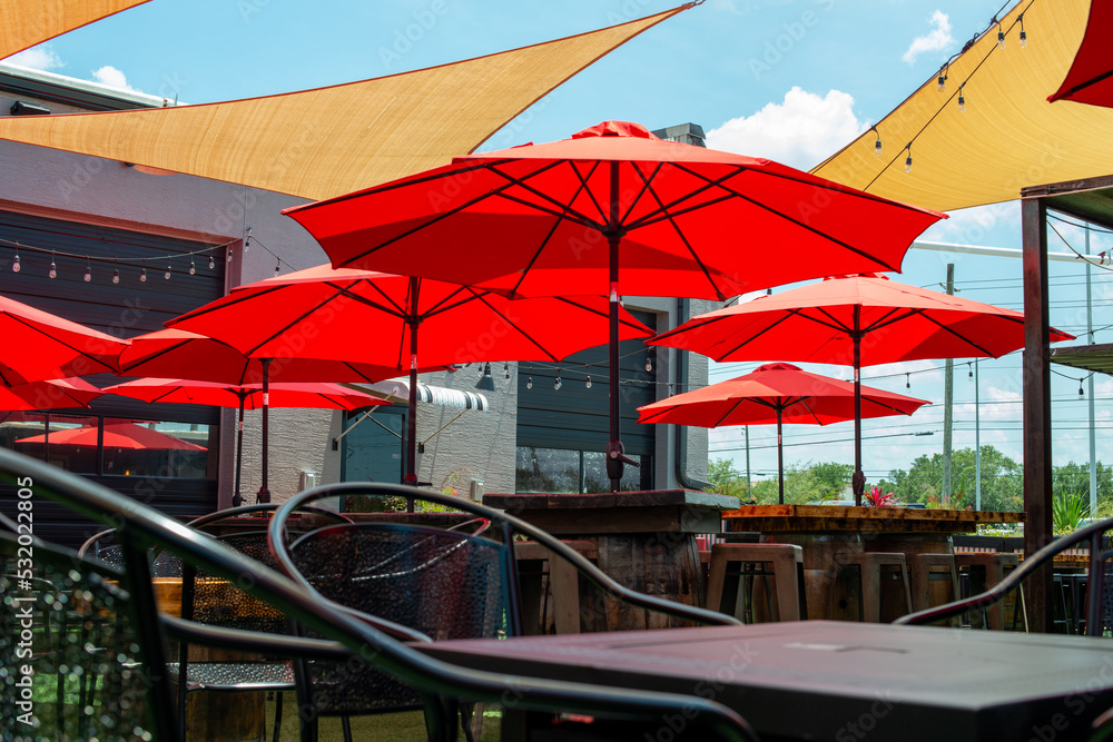 Fototapeta premium Multiple triangle shaped yellow nylon sunshades and awnings hanging over a patio deck. There are red colored canvas umbrellas hung with strings of clear patio light against a bright blue sunny sky.
