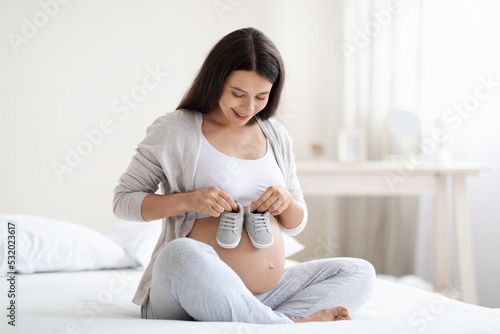 Cheerful pregnant woman holding baby shoes on big tummy
