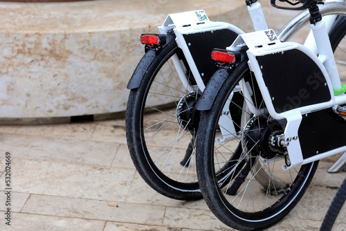 Rental E-bikes, back view of two bicycles assisted by electric motors with QR Coder, are parked in a row in the city street. Modern rental electric bikes standing a row during charging. European eco.