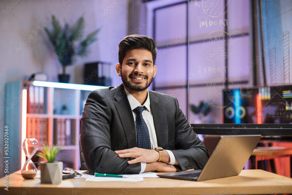 Fototapeta premium Front view portrait of young smiling experienced smart stylish bearded hindu office manager in suit sitting at table using laptop in evening office.