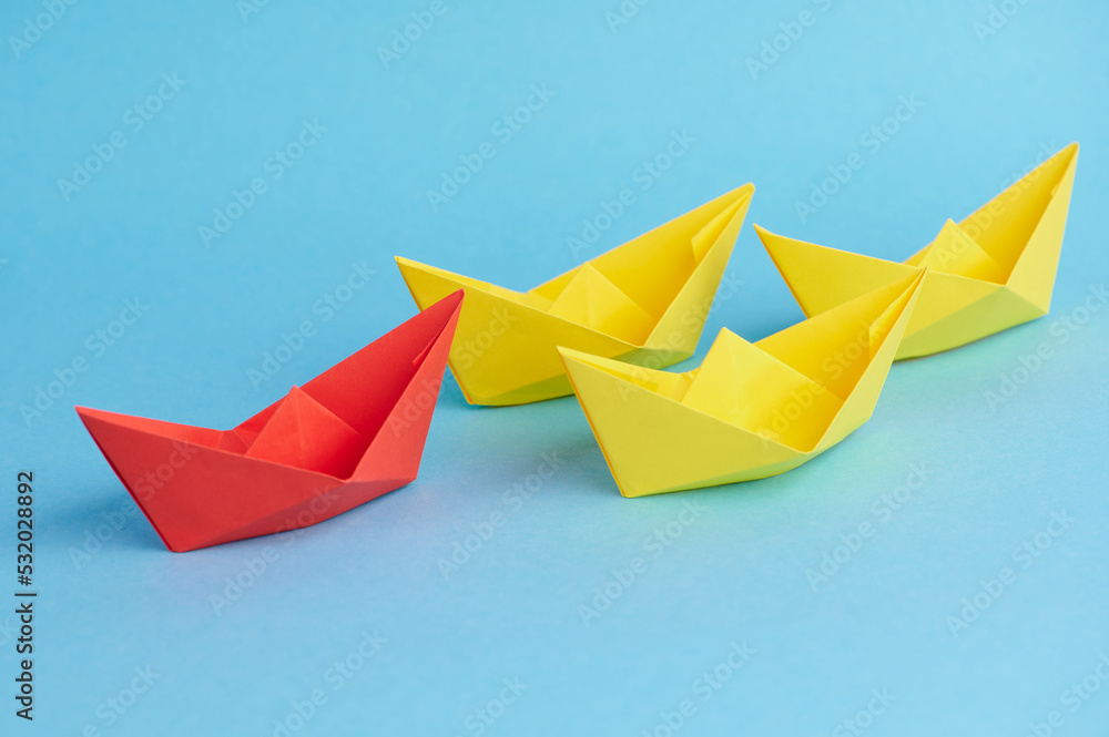 A red paper boat leads small yellow paper boats as a team. Concept for teamwork, cooperation, leadership as well as partnership. A red paper boat leads small yellow paper boats as a team. Concept for teamwork, cooperation, leadership as well as partnership.